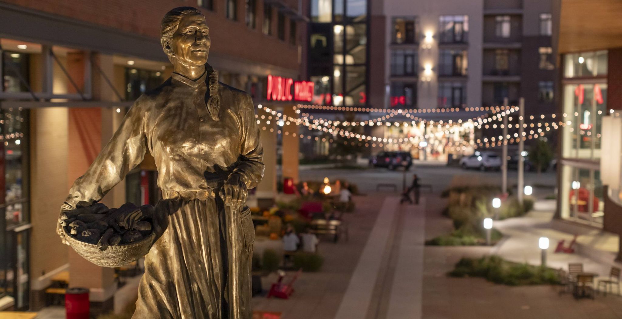 Lenexa Public Market in Kansas view of street with bronze statue and string lights at dusk